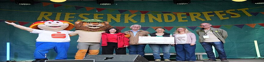 Spendenscheckübergabe mit Maskottchen von Sparkasse und Zoo auf der Bühne 